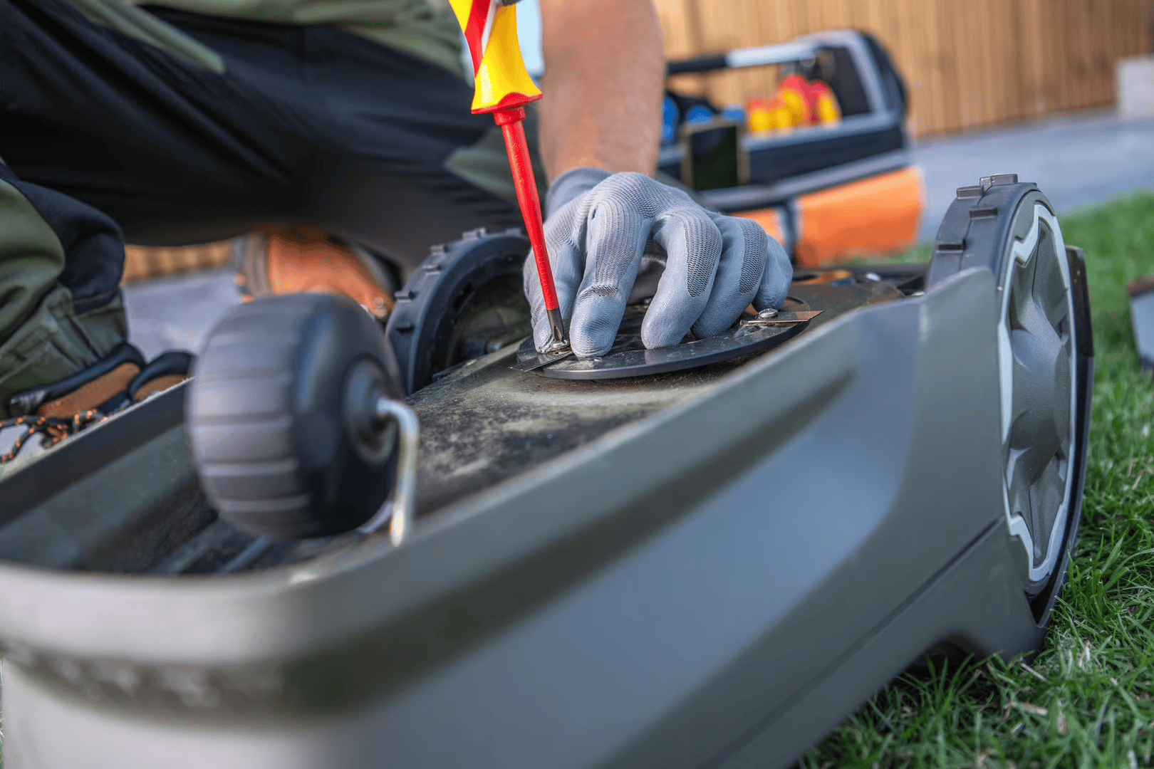 Person in gloves using a screwdriver to work on a robotic lawnmower in a grassy yard.