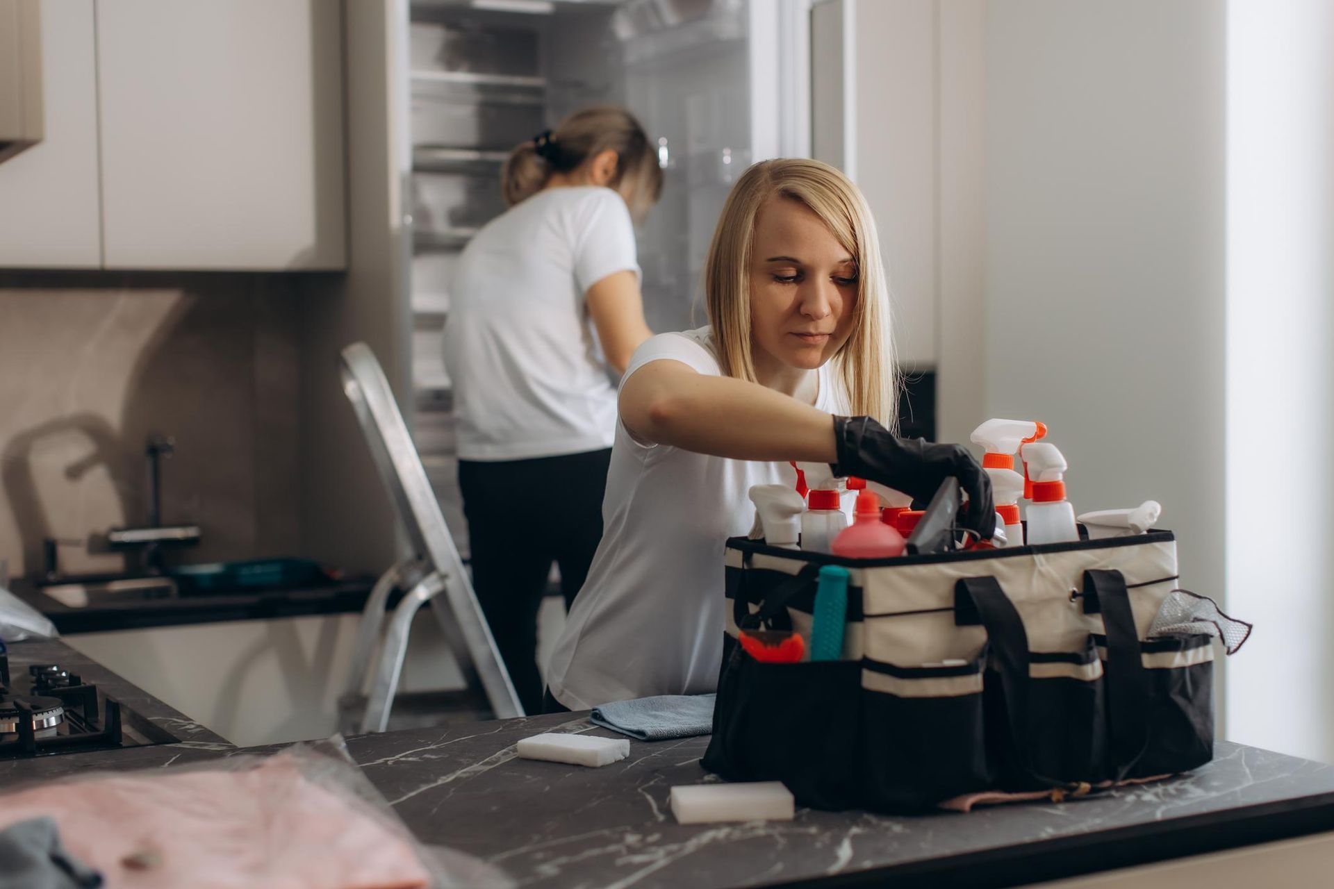 Woman wearing yellow gloves cleaning floor in a living room, smiling. Cleaning supplies in a blue bin.