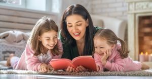 A woman is reading a book to two little girls while laying on the floor.