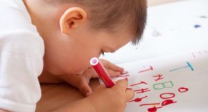 A young boy is writing on a piece of paper with a red marker.