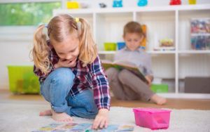 A little girl is playing with a puzzle on the floor while a boy sits on the floor reading a book.