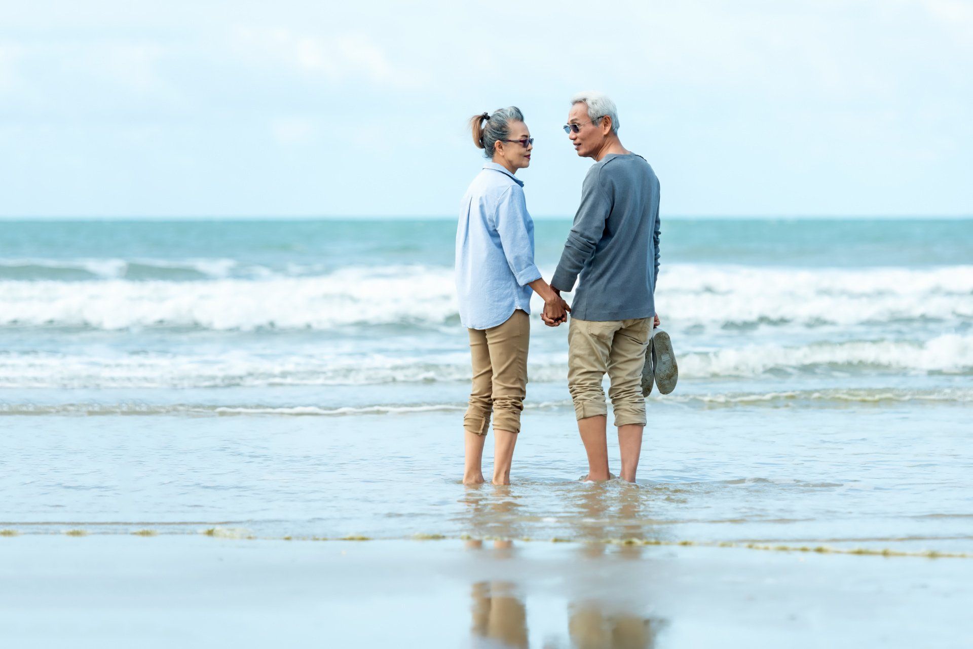 An elderly couple is standing on the beach holding hands.