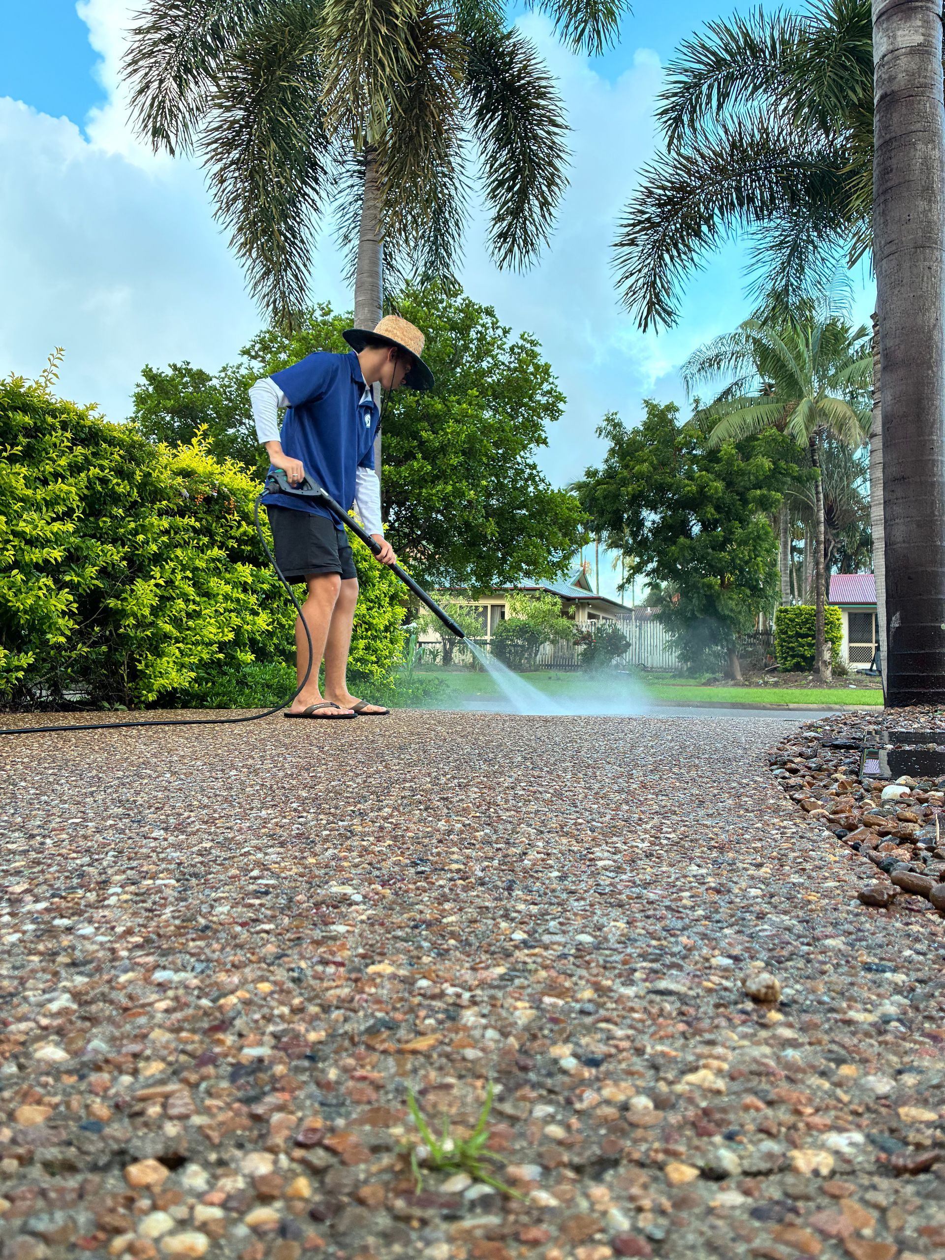 A Person in A Sun Hat Uses a Pressure Washer to Clean a Textured Pebble Driveway — Schmucker Cleaning In Gulliver, QLD