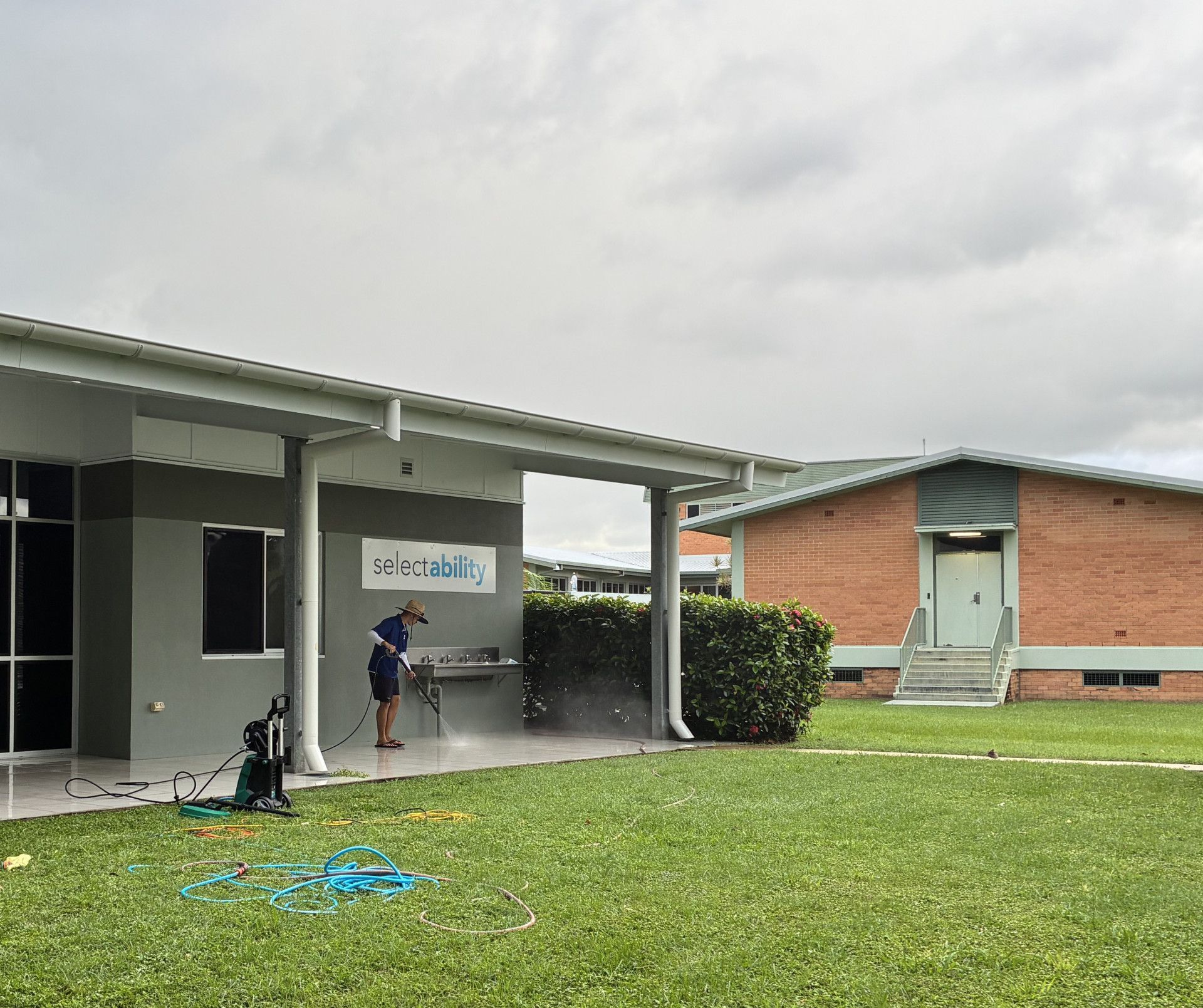 Person Uses a Pressure Washer to Clean a Paved Patio Area Outside a Grey Building — Schmucker Cleaning in Gulliver, QLD