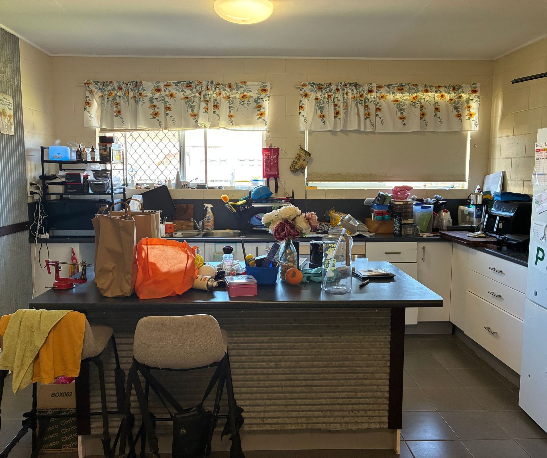 Cluttered Kitchen With Island And Two Windows With Floral Valances — Schmucker Cleaning in Gulliver, QLD