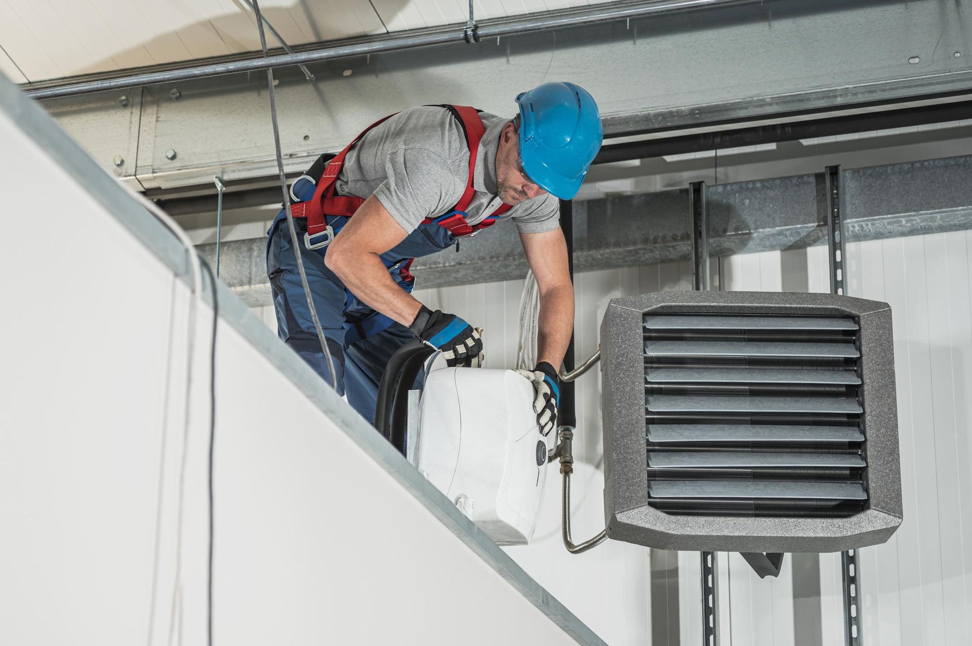A worker in a blue hard hat and safety harness installs a heating unit on a ceiling.