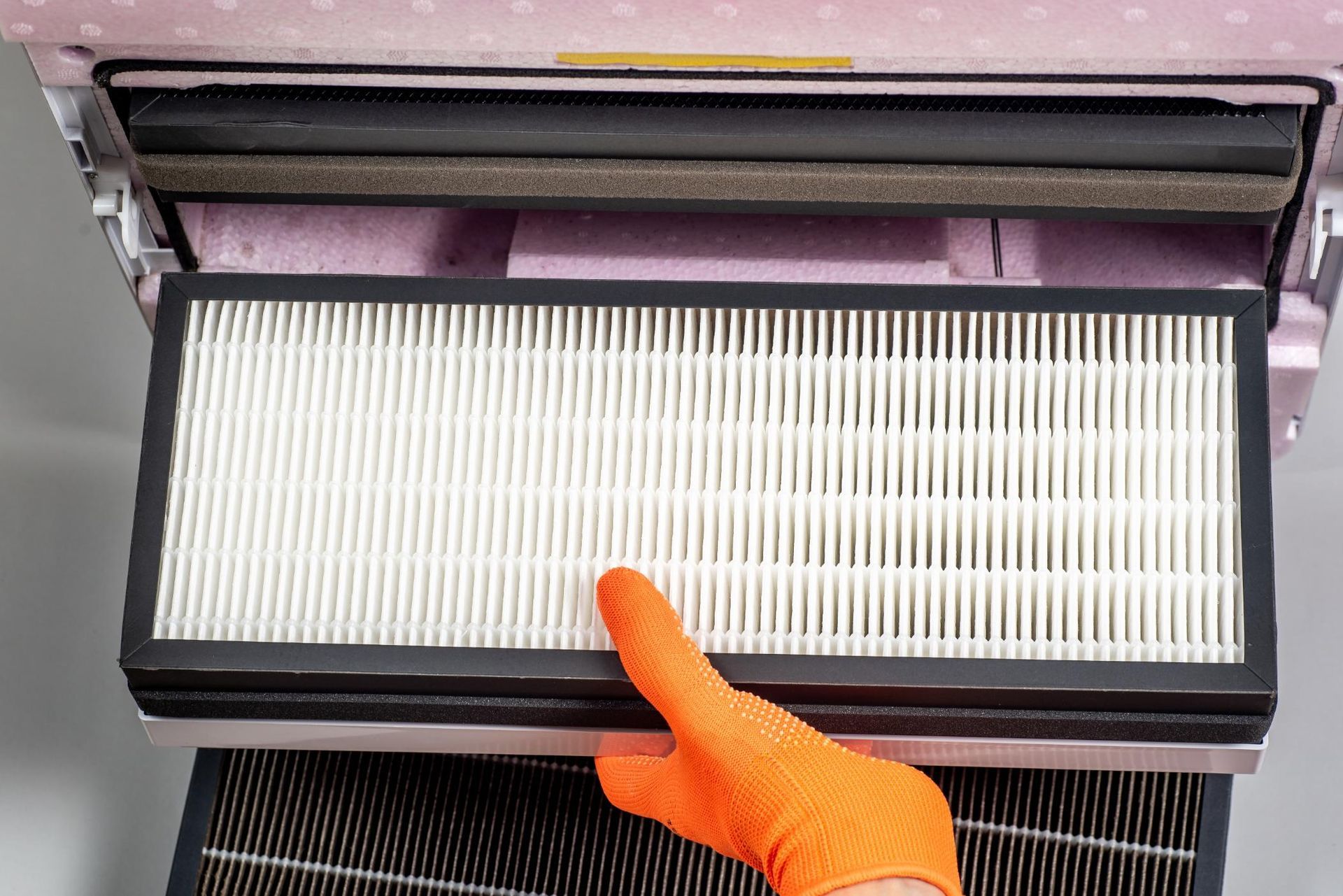Hand in orange glove holding air filter from pink air purifier.