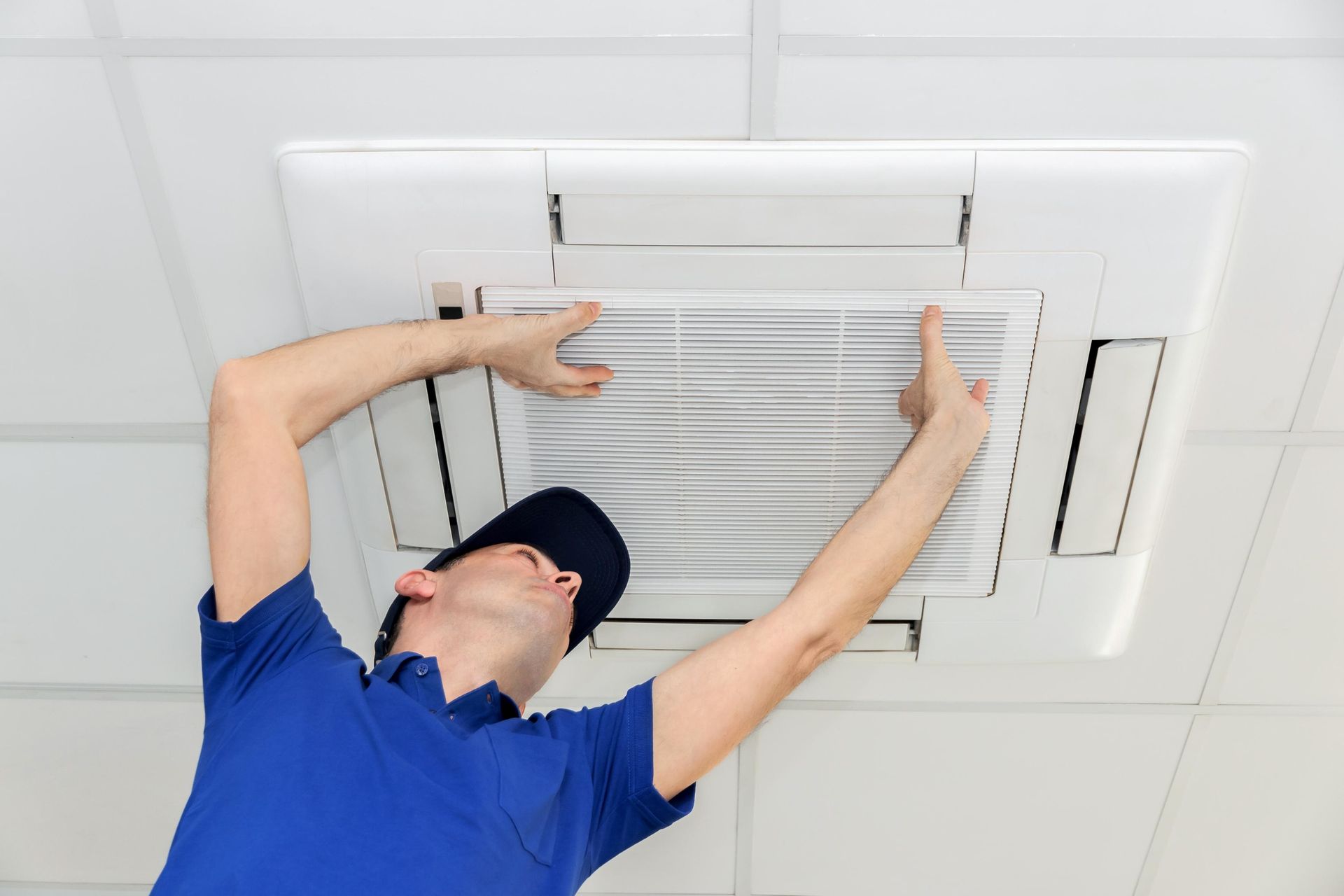 Man in blue shirt and cap installing an air filter in a ceiling-mounted unit.