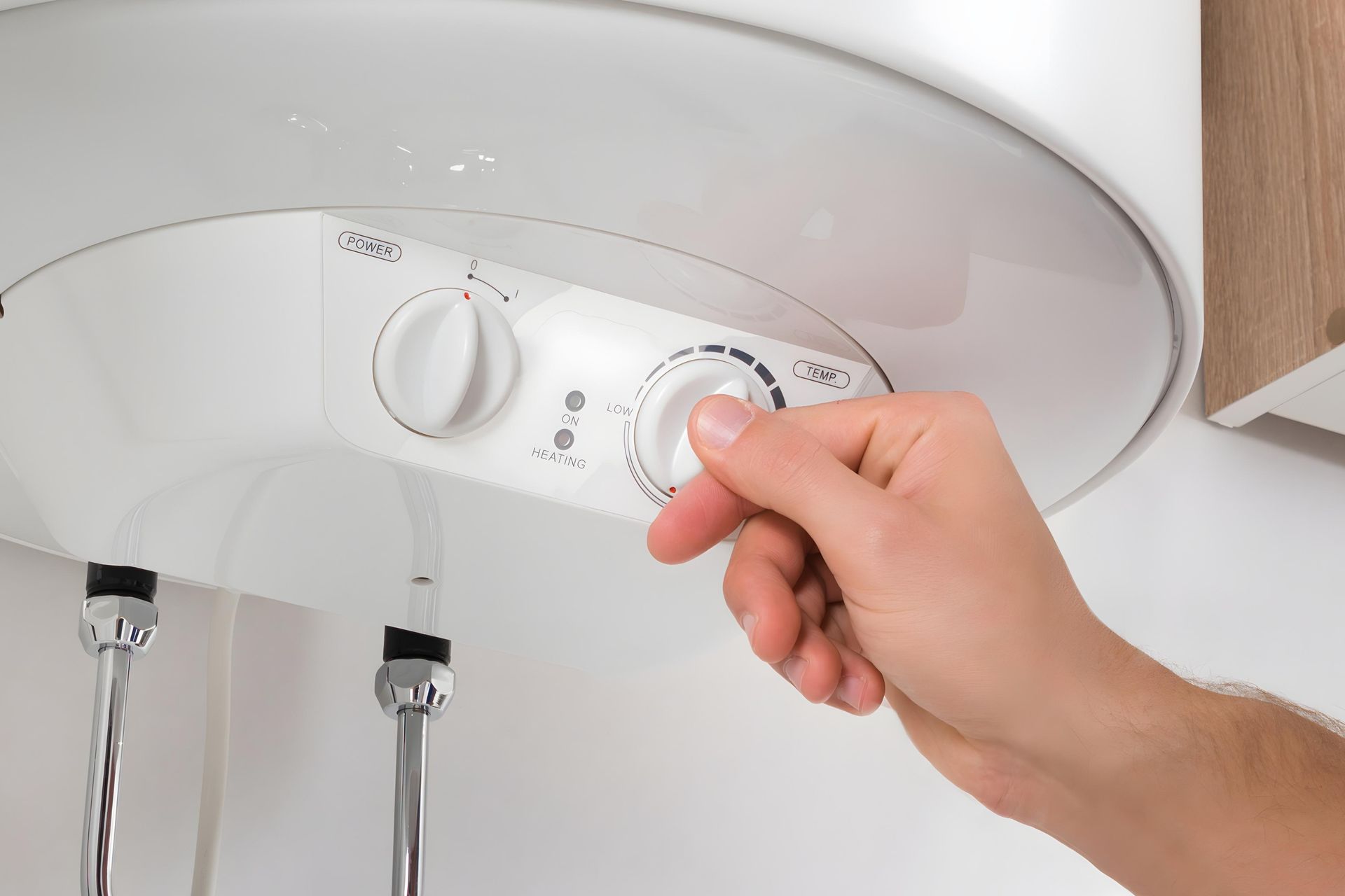 Hand turning a dial on a white water heater, next to silver pipes, in a white room.