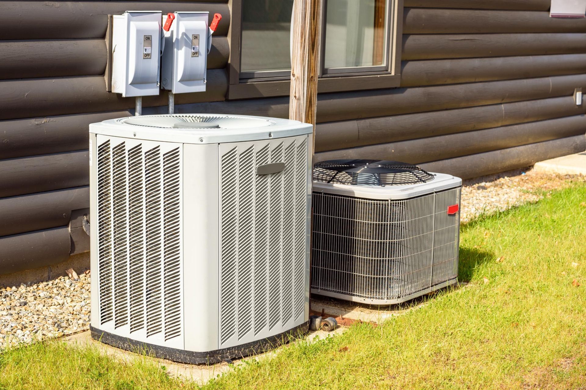 Two air conditioning units beside a dark brown house, with electrical boxes overhead.