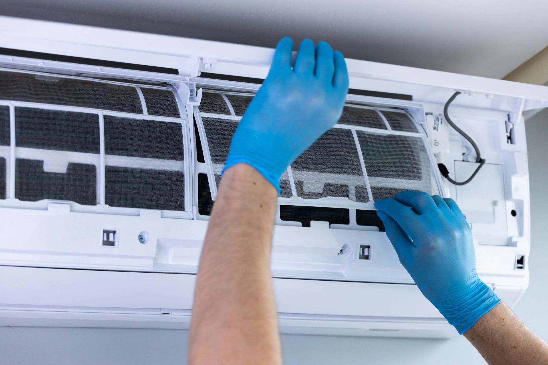 Person in blue gloves cleaning the air filter of a white air conditioning unit.