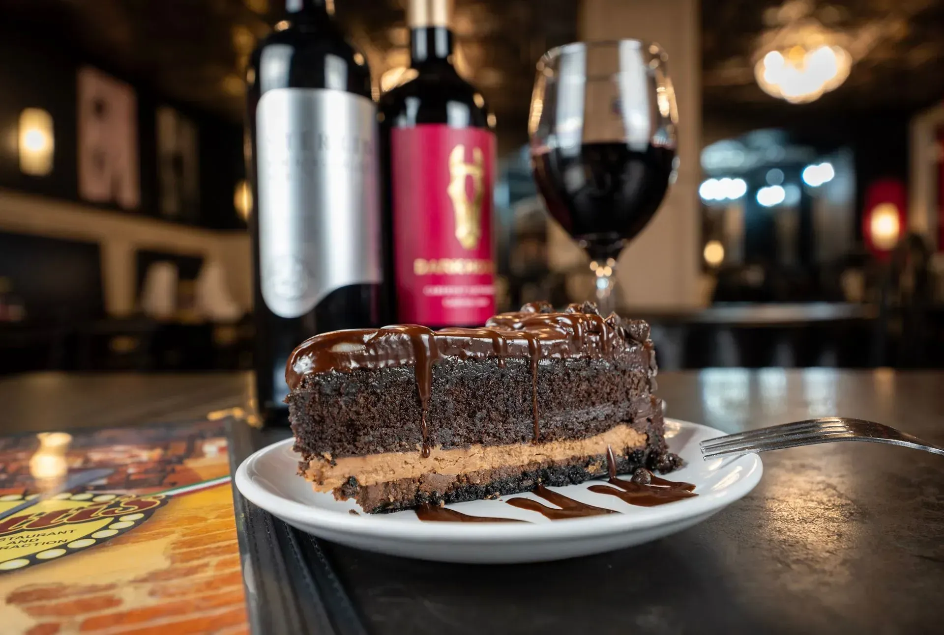 A piece of chocolate cake is on a plate next to a glass of wine and a bottle of wine.