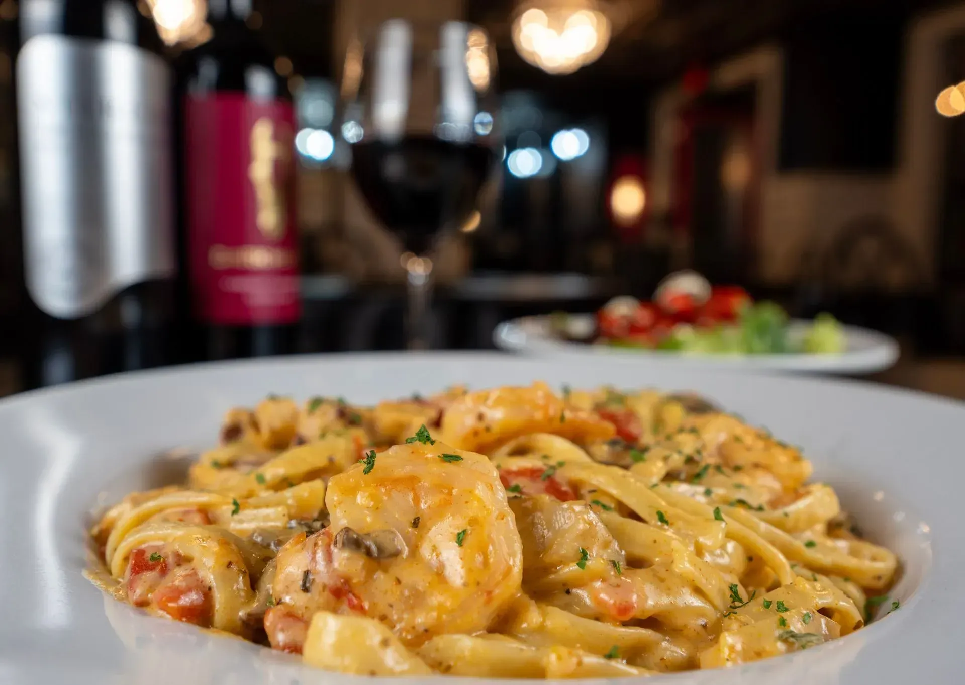 A plate of pasta with shrimp and a glass of wine in the background.
