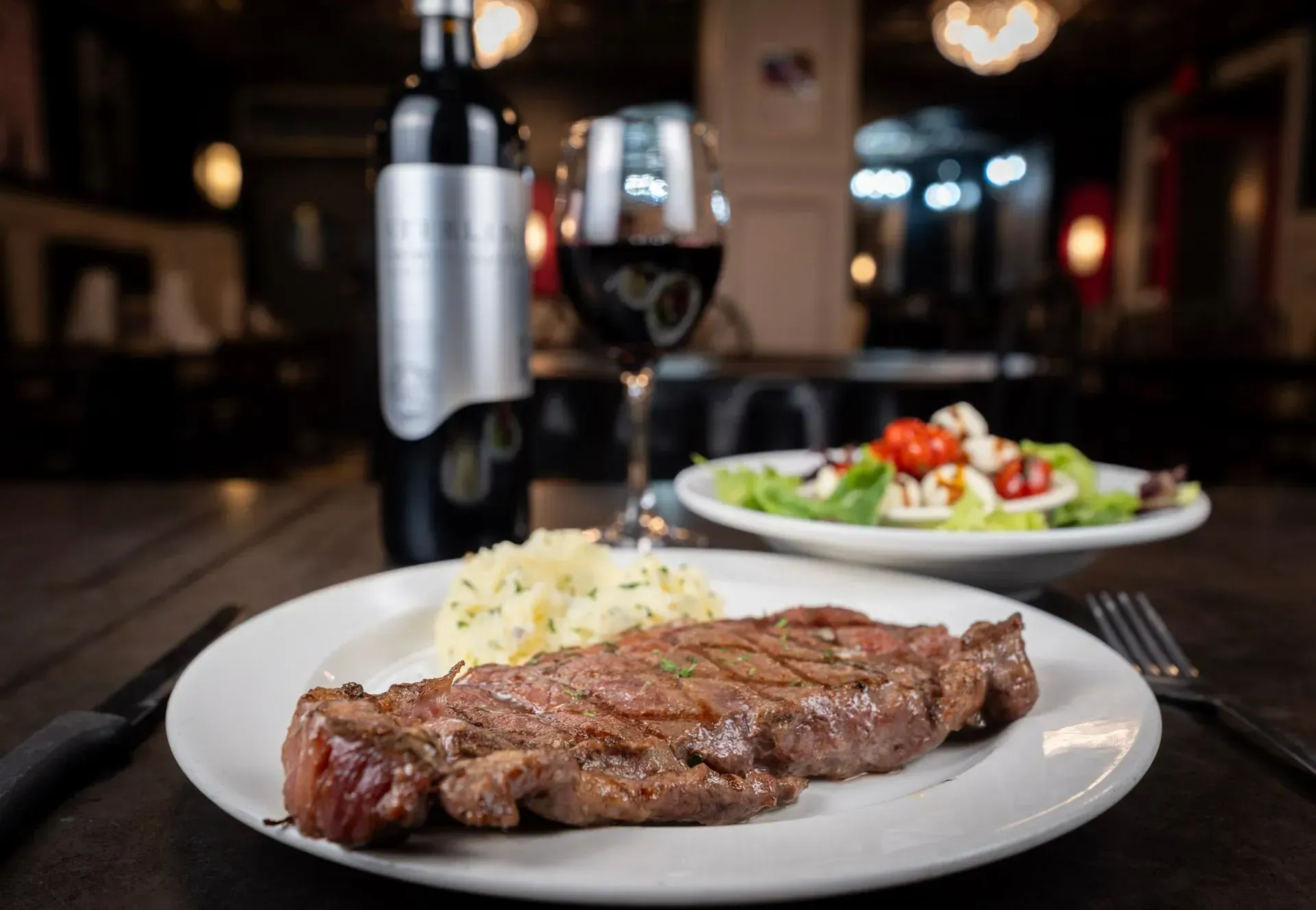 A white plate topped with a steak and mashed potatoes next to a bottle of wine and a glass of wine.