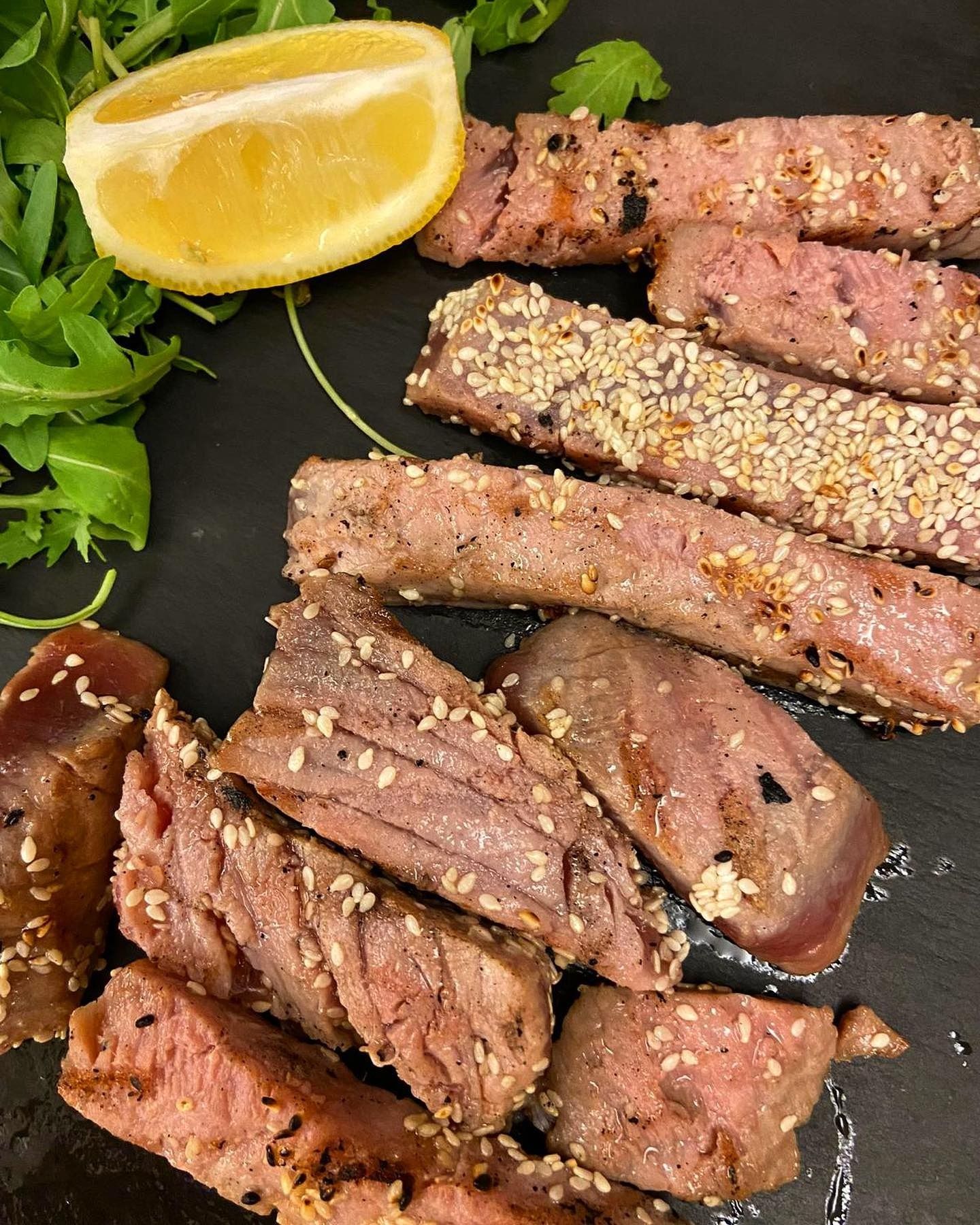 Fette di tonno scottato con semi di sesamo, spicchio di limone e verdure su una superficie scura.