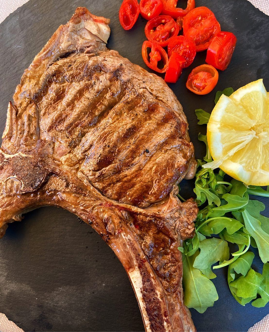 Bistecca alla griglia con pomodori, limone e rucola su un piatto nero.