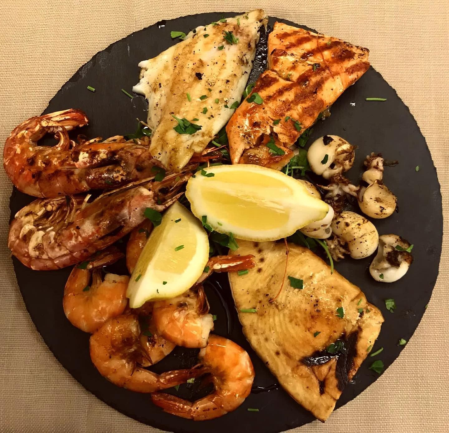 Piatto di frutti di mare alla griglia: gamberi, filetti di pesce, calamari, spicchi di limone, su un piatto nero.