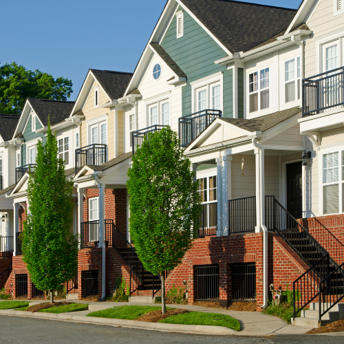 Row of townhouses with varying colors, brick bases, and small trees in front.