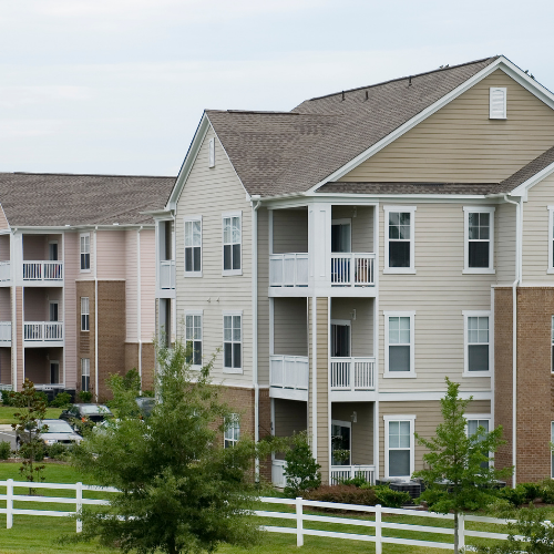 Multi-story apartment buildings with beige and brick siding, white balconies, and a white fence in front.