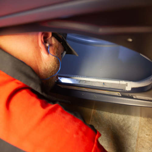 Man in orange jumpsuit inspects a dryer. He wears glasses and a hat, working near the open door in an indoor setting.