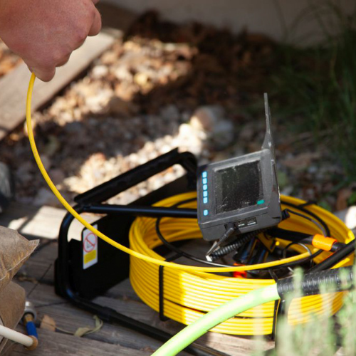Person using a yellow sewer inspection camera. The setup includes a coiled cable and a screen in an outdoor setting.