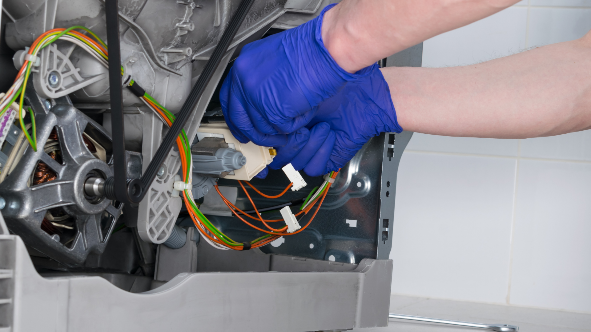 Hands in blue gloves connect wires inside a washing machine, likely for repair.