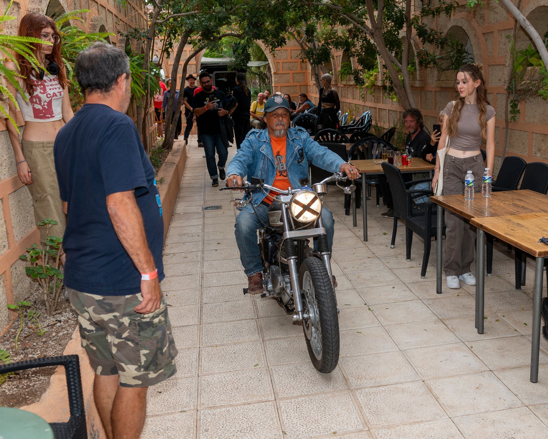 A man is riding a motorcycle down a sidewalk with tables and chairs in the background