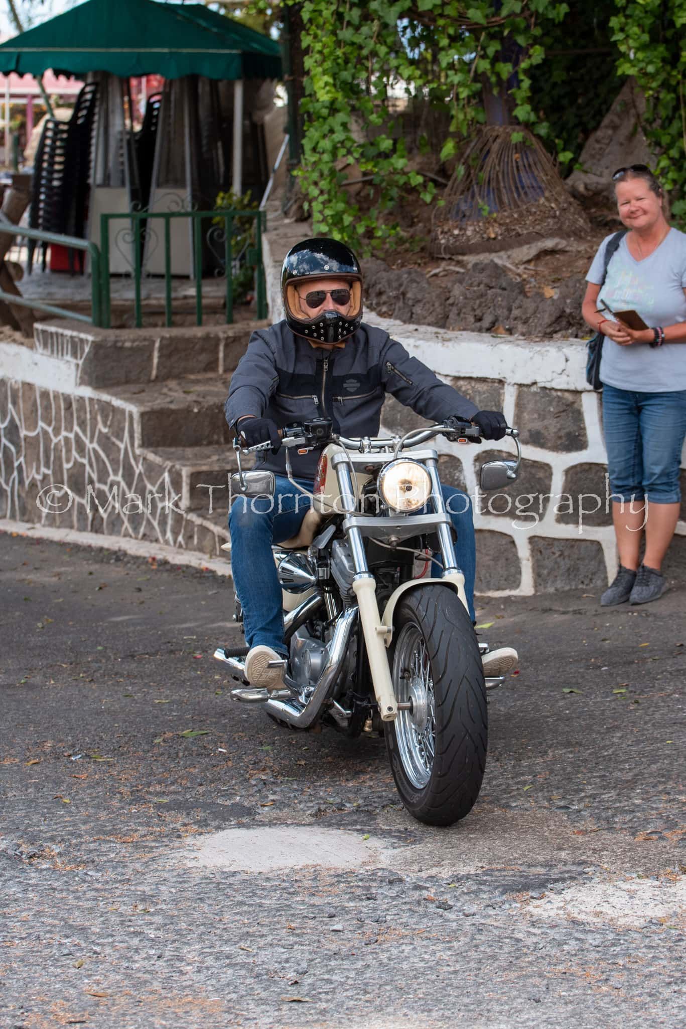 A man is riding a motorcycle down a street while a woman looks on.