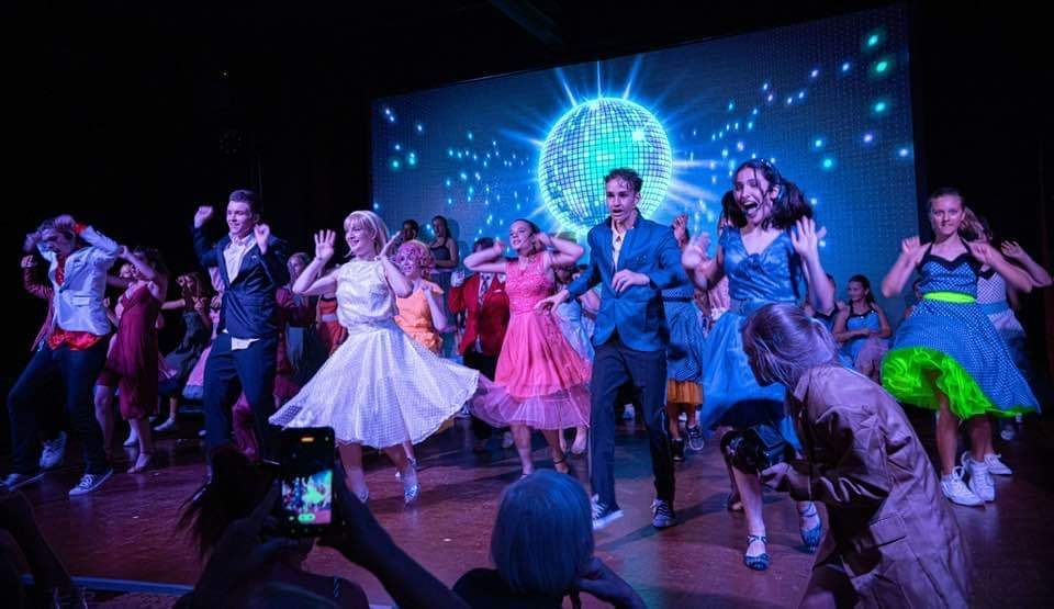 A group of people dancing on a stage with a disco ball background. Some people are holding up phones.
