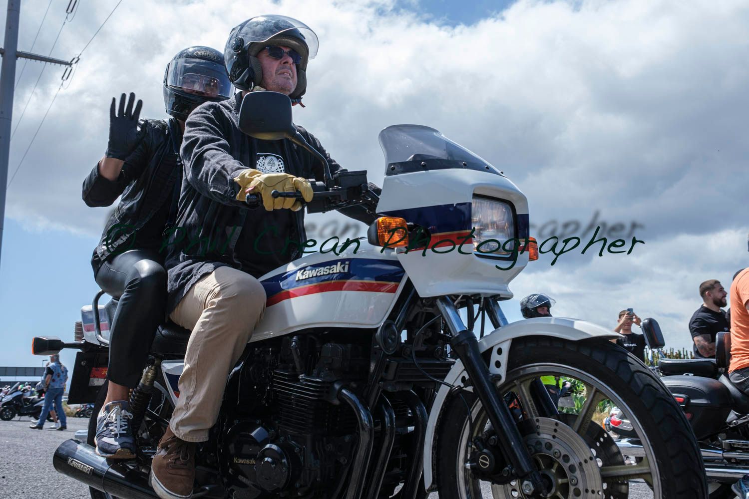 A man and a woman are riding a motorcycle and waving at the camera.