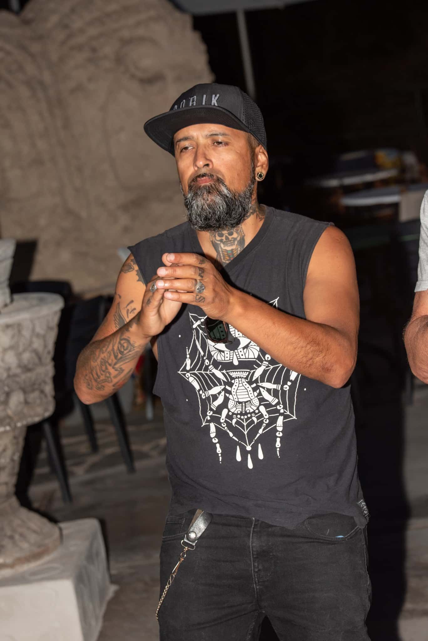 A man with a beard and tattoos is wearing a hat and a black tank top.