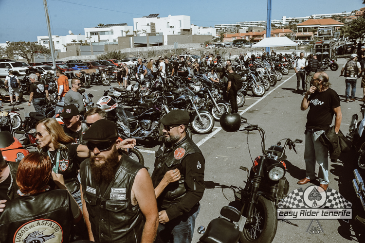 A group of people are standing in front of a parking lot filled with motorcycles.