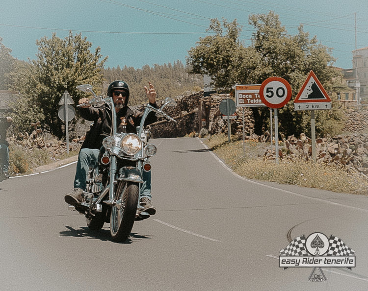 Man riding a motorcycle on a road, raising his arms. Speed limit sign of 50 km/h is visible.