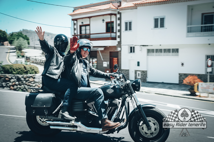 Two people on a Harley Davidson motorcycle wave; white building in the background.