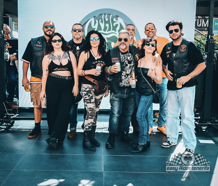 Group of people, possibly a motorcycle club, posing outdoors. Most wear black vests, hold drinks, and are smiling.
