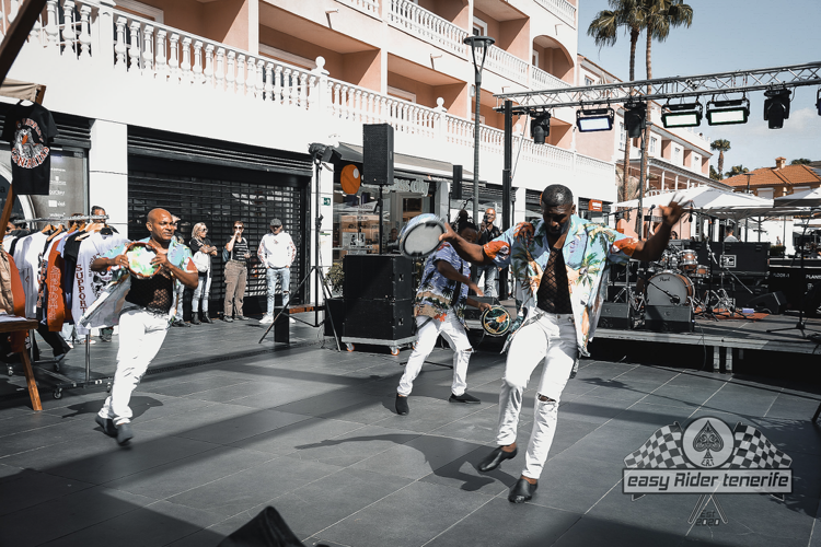 A band performing outdoors. Three people dancing on a stage in front of a building with a merchandise stand.