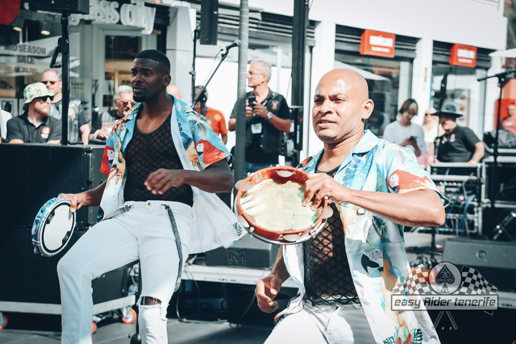 Two men playing tambourines on a stage. They wear white pants, patterned shirts and fishnet undershirts.