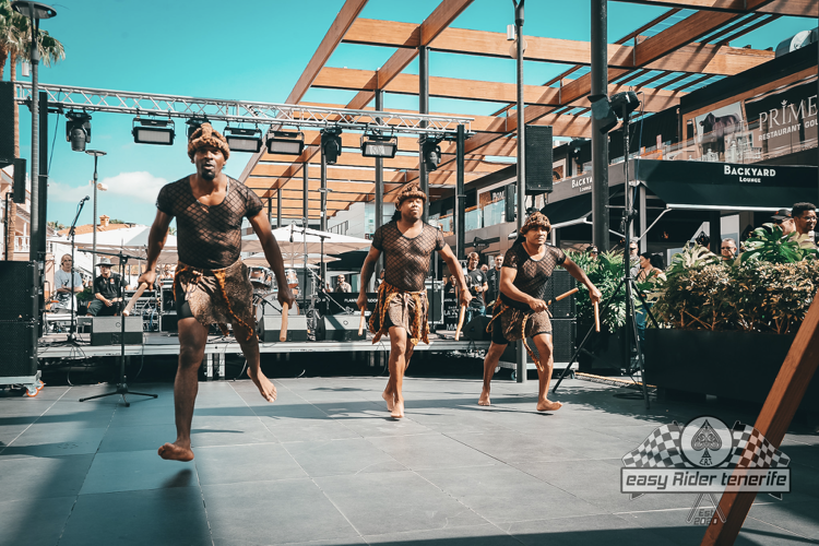 Three dancers in brown costumes performing on an outdoor stage.