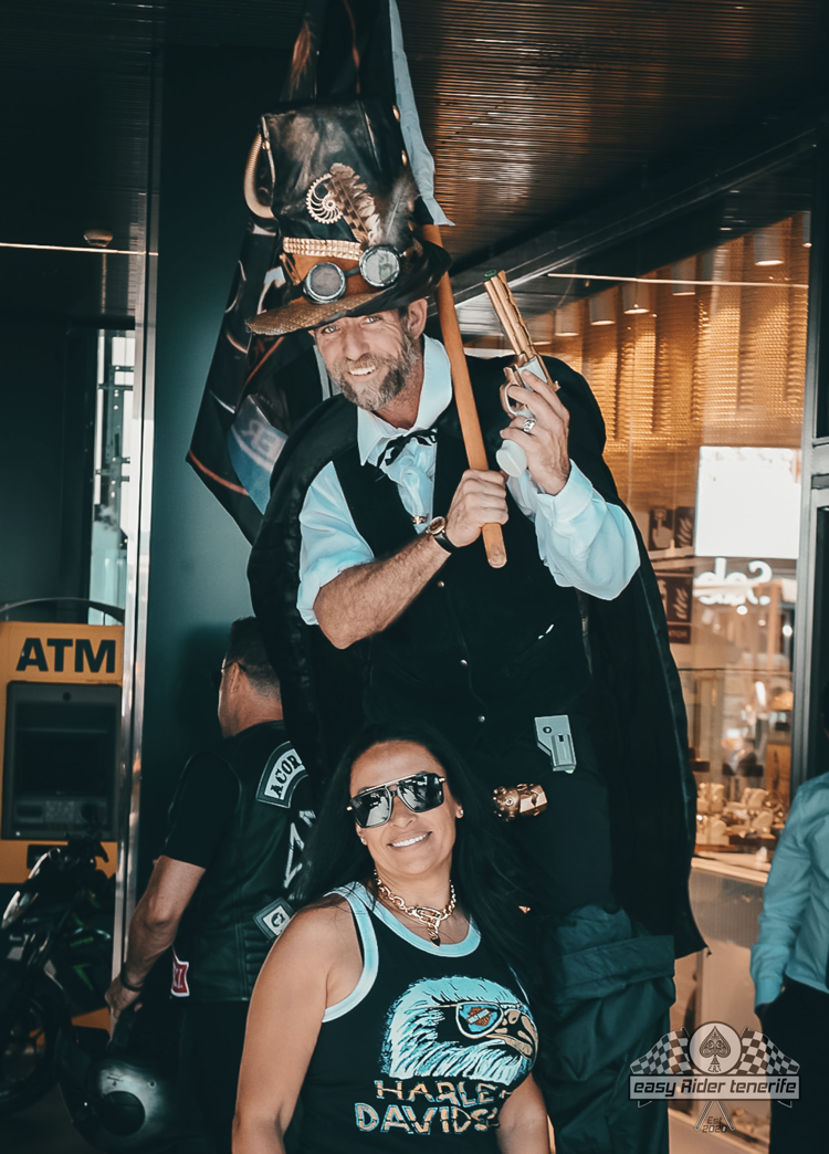 A person on stilts in steampunk attire holds wooden guns, smiling, with a woman in a Harley-Davidson tank top.