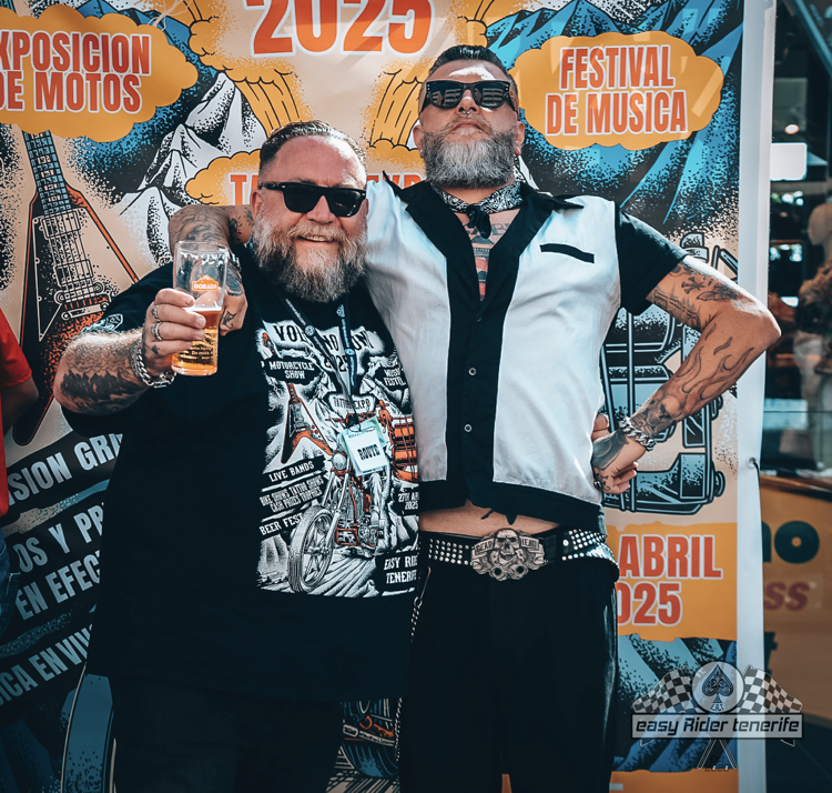 Two men pose smiling in front of a festival banner, one holding a beer.
