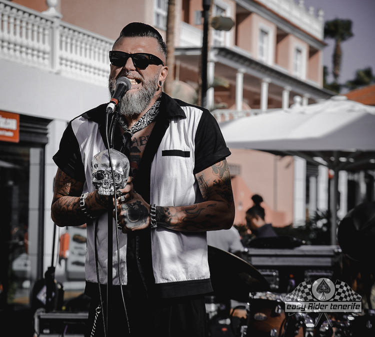 Man with tattoos singing into a microphone, holding skull guitar, in outdoor setting.