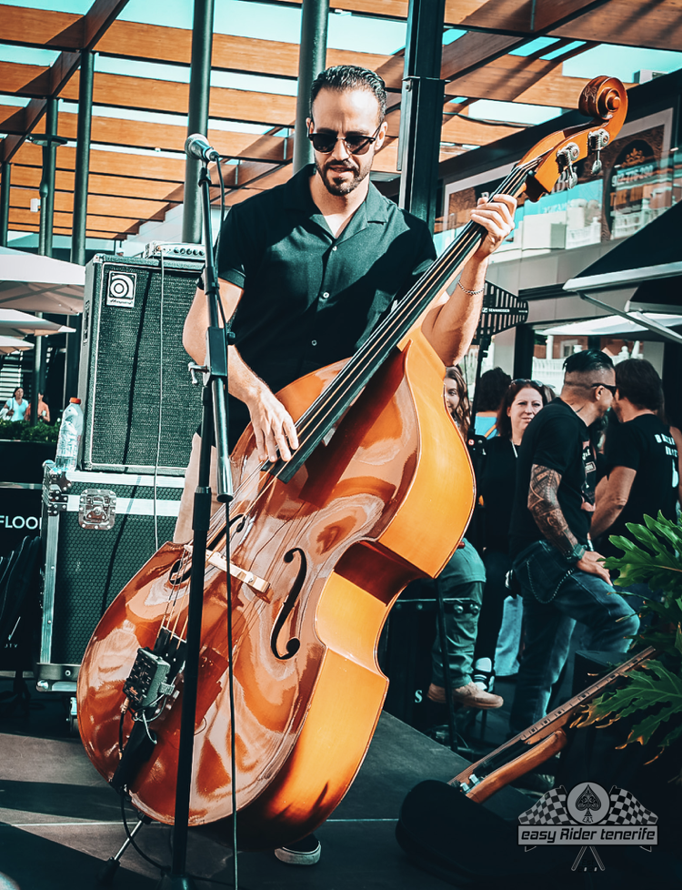 Man playing a stand-up bass guitar on a stage outside. He wears a black shirt and sunglasses. Other people are in the background.