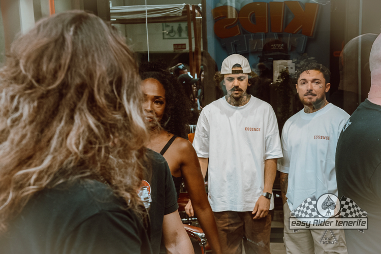 Group of people outside a shop, one person in focus with long hair, others in white shirts, a 