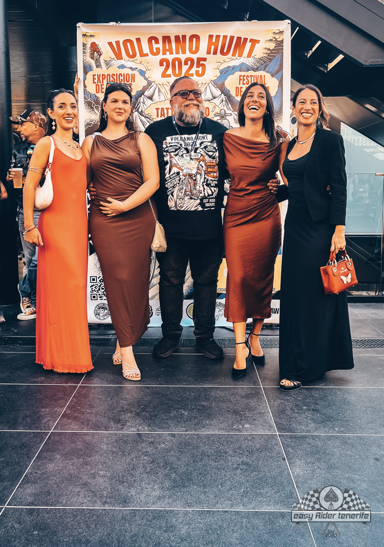 Five people pose for a photo in front of a sign. One wears an orange dress, and three wear brown dresses. A man in the center wears a graphic t-shirt.