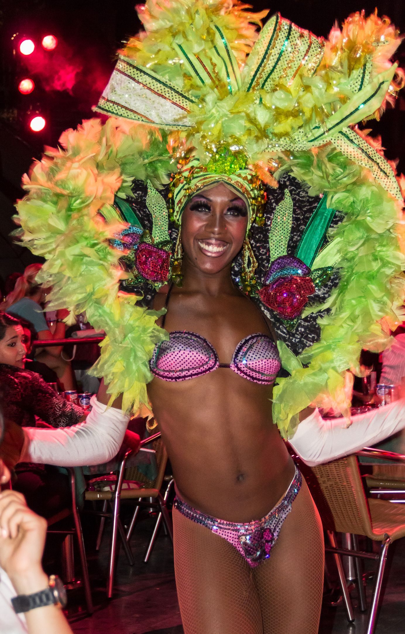 Woman in elaborate headdress and bikini, smiling, posing in a restaurant with vibrant green and pink details.