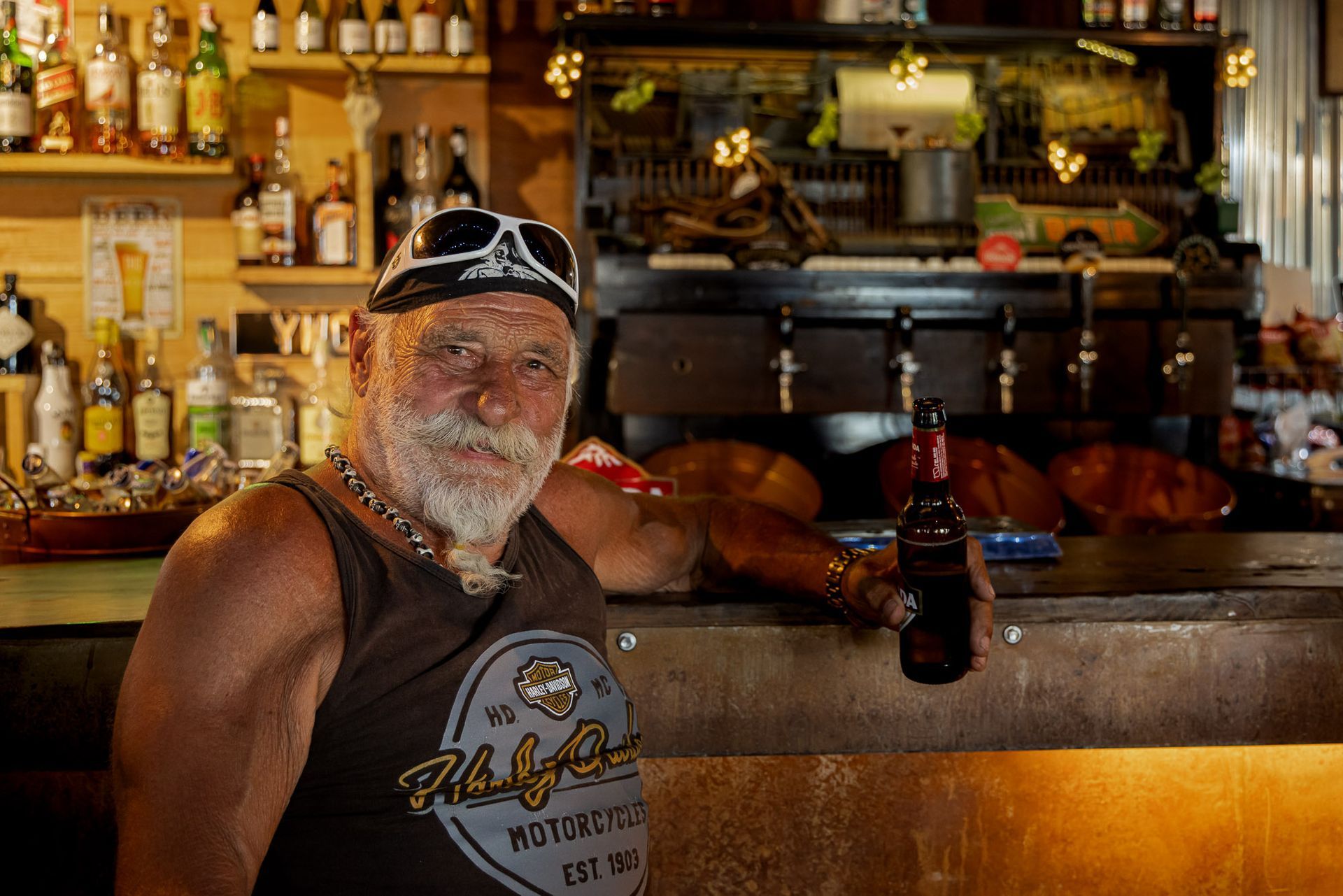 A man with a beard is sitting at a bar holding a bottle of beer.