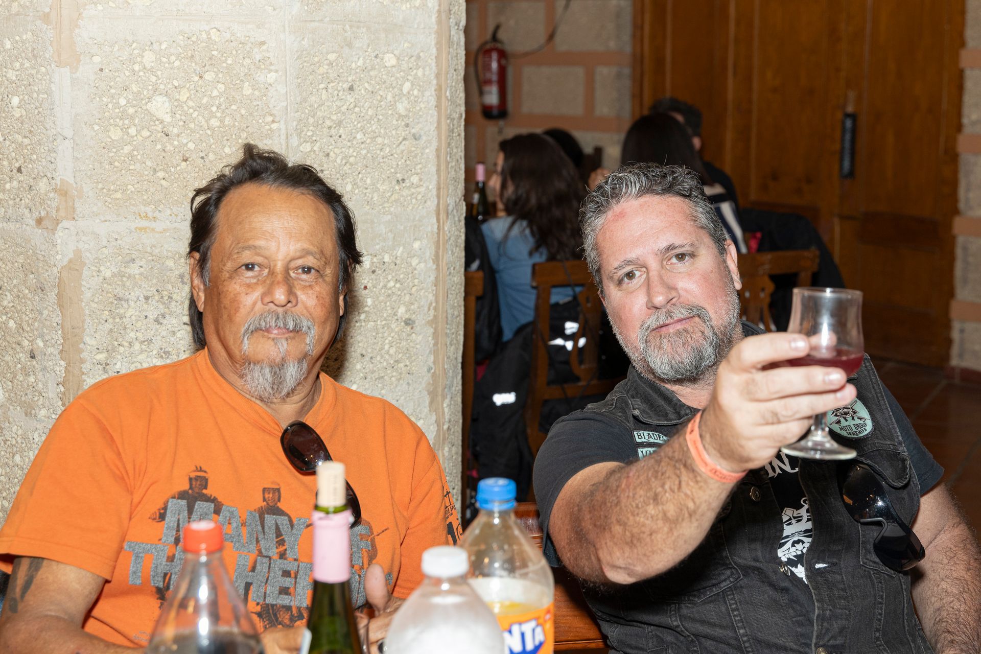 Two men are sitting at a table with bottles of fanta and a glass of wine.