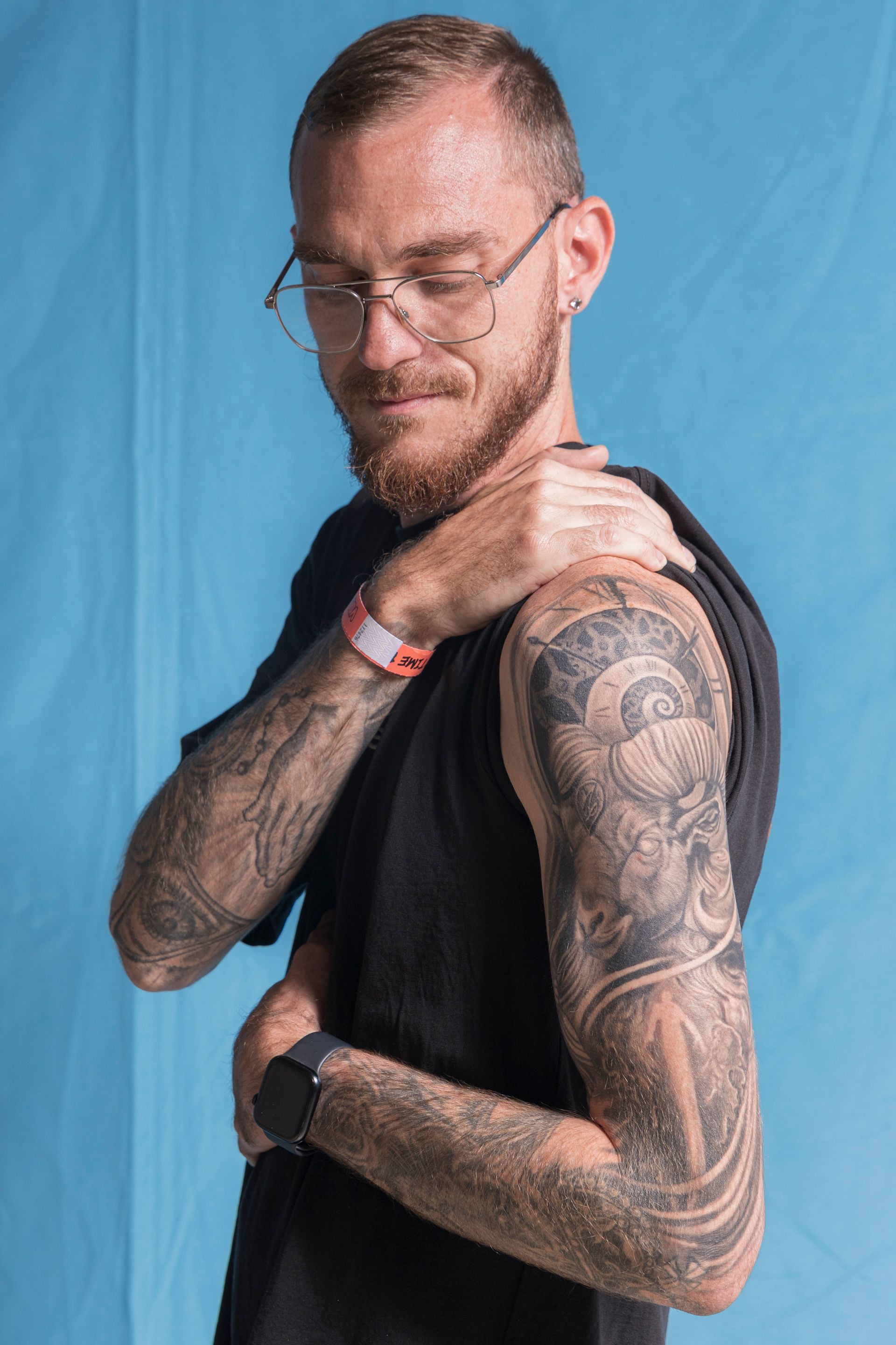 A man with glasses and a tattoo on his arm is holding his shoulder.