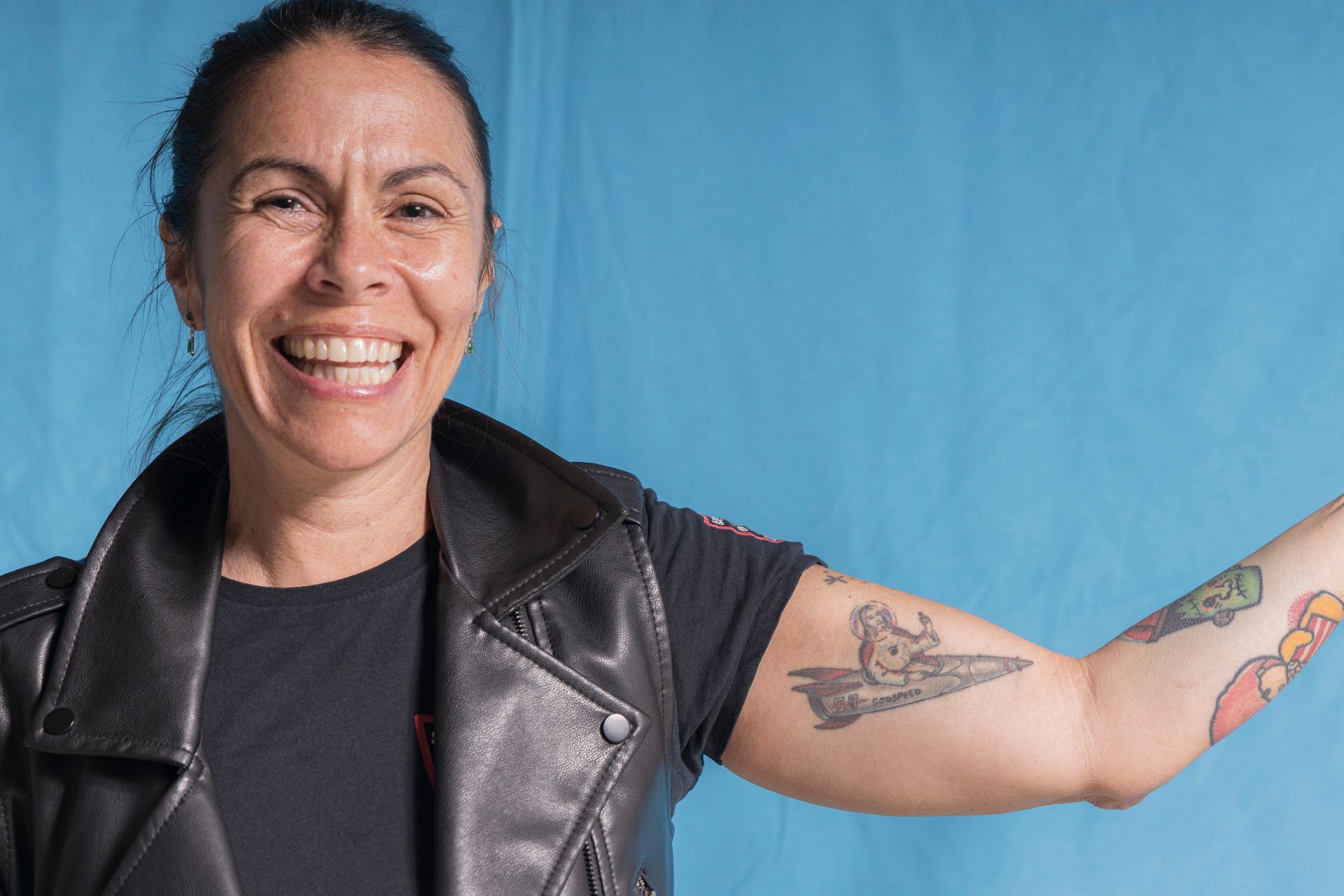 A woman with a tattoo on her arm is smiling and wearing a leather vest.