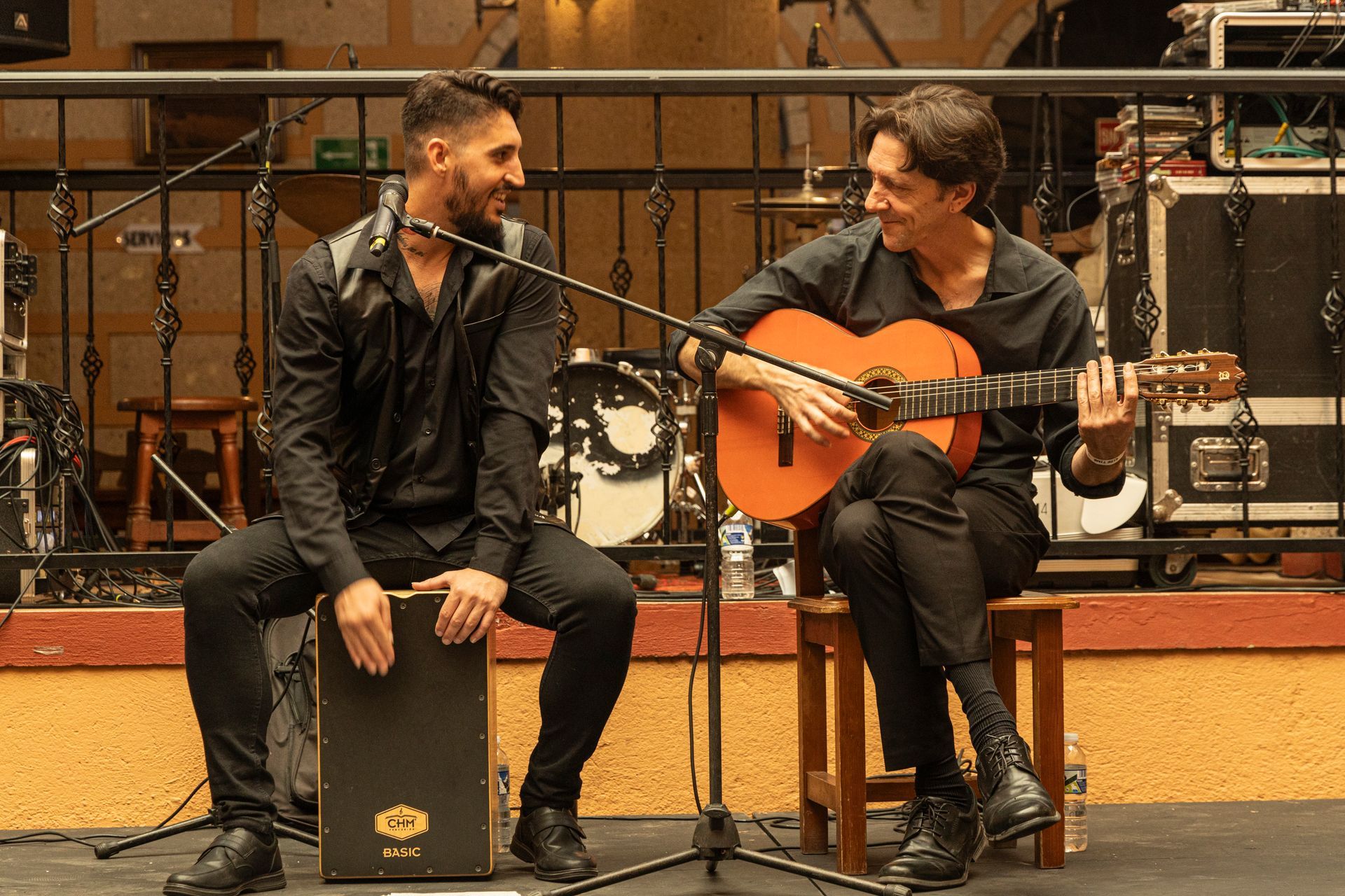 Two men are sitting on stools playing guitars and singing into microphones.